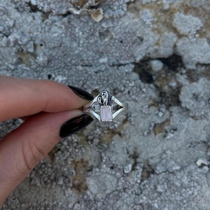 May include: A silver ring featuring a rectangular, pale pink gemstone. The ring has a decorative design on the band and around the gemstone. The ring is held against a textured, grey background.