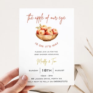 May include: A white baby shower invitation with a watercolor illustration of a bowl of red apples. The text reads "The apple of our eye is on it's way."  The invitation includes the names "Madly & Tim", the date "Sunday 18th August", the time "10am - 3pm", the location "88 Lagoon Drive, Perth WA", and the RSVP information "Kindly RSVP to Polly on 0453627272".