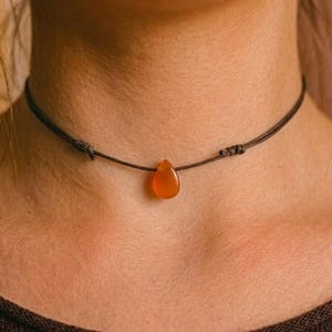 Może przedstawiać: Brązowy naszyjnik typu choker z wisiorkiem w kształcie łzy z pomarańczowego kamienia szlachetnego. Wisiorek zawieszony jest na cienkim, wiązanym sznurku. Naszyjnik jest noszony na szyi.