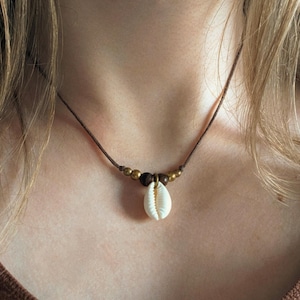 Könnte beinhalten: Eine braune Kordelkette mit einem weißen Muschelanhänger. Die Halskette hat kleine, runde, goldfarbene Perlen und eine dunkelbraune Perle in der Nähe der Muschel. Die Halskette wird um den Hals getragen.