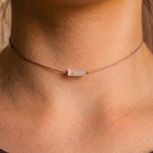 Może przedstawiać: Delikatny naszyjnik typu choker z małą, prostokątną zawieszką z jasnego kamienia szlachetnego. Zawieszka zawieszona jest na cienkim, brązowym sznurku, tworząc minimalistyczny i elegancki design. Naszyjnik noszony jest na szyi.