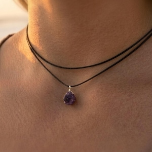 Könnte beinhalten: Eine Halskette mit doppeltem schwarzem Band und einem einzelnen, rohen, tiefvioletten Amethyst-Anhänger, der an einer silberfarbenen Drahtwicklung hängt. Die Kette liegt am Hals an und betont den Kontrast zwischen dem dunklen Band und dem Edelstein.