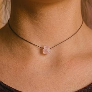 Può includere: Una delicata collana girocollo con un pendente in quarzo rosa grezzo. La gemma rosa pallido è sospesa a un sottile cordino marrone scuro. La collana mette in risalto la bellezza naturale del cristallo. Il pendente ha una forma irregolare.