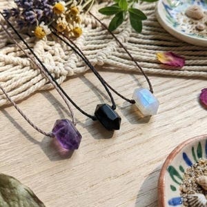 May include: Three gemstone necklaces with black and tan cords. The gemstones are amethyst, black onyx, and moonstone. The necklaces are displayed on a wooden surface with decorative elements.