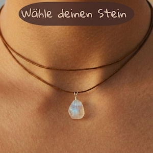 Könnte beinhalten: Eine zweireihige braune Kordelkette mit einem einzelnen, rohen, schillernden Mondstein-Anhänger, der mit Silberdraht umwickelt ist. Der Text "Wähle deinen Stein" steht in einer dunkelbraunen ovalen Form oben im Bild.