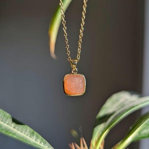 Könnte beinhalten: Eine goldfarbene Halskette mit einem quadratischen, pfirsichfarbenen Anhänger. Der Anhänger ist in einen goldenen Rahmen gefasst und hängt an einer zarten Goldkette. Der Hintergrund ist dunkelgrau, mit grünen Blättern im Vordergrund.