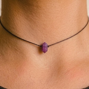 May include: A choker necklace with a small, purple gemstone pendant. The gemstone is faceted and hangs from a thin, dark brown cord. The necklace is worn around the neck.