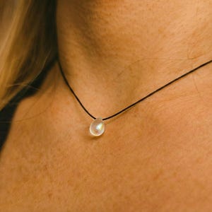 Può includere: Una delicata collana con un piccolo pendente iridescente a forma di goccia sospeso a un sottile cordino nero. Il pendente sembra essere fatto di un materiale traslucido, che riflette la luce. La collana è indossata vicino al collo.