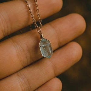 Puede incluir: Un delicado collar con un colgante de cristal azul pálido. El cristal está envuelto en alambre de metal dorado y cuelga de una cadena a juego. El fondo es una superficie borrosa de tonos cálidos.