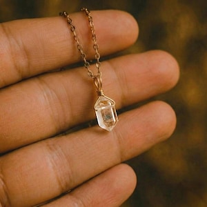 Op de afbeelding: Een delicate goudkleurige ketting met een hanger van heldere kwartskristal. De kristal is facetgeslepen en hangt aan een fijne ketting. De hanger is omwikkeld met goudkleurig draad, wat een vleugje elegantie toevoegt aan dit sieraad.