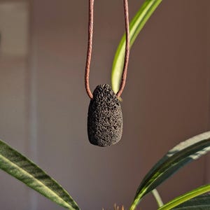 Könnte beinhalten: Ein schwarzer Lavastein-Anhänger an einer braunen Lederschnur. Der Anhänger ist ein natürlicher Stein mit rauer Textur.