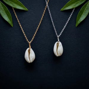 Puede incluir: Dos collares de conchas marinas sobre un fondo negro. Uno tiene una cadena dorada y el otro una cadena plateada. Cada collar presenta un colgante de concha de cauri blanca. Hojas verdes en las esquinas superiores.