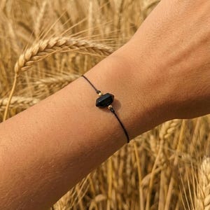 Op de afbeelding: Een armband met een zwarte edelsteen en gouden details. De armband heeft een donkere, facetgeslepen steen in het midden, geregen aan een dun zwart koord. Kleine gouden kralen zijn aan weerszijden van de edelsteen geplaatst. De achtergrond is een tarweveld.