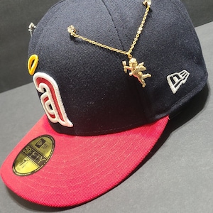 May include: A navy blue baseball cap with a red brim, featuring a gold cherub pendant and a white logo. The cap has a 59FIFTY sticker and a team logo with a white outline.