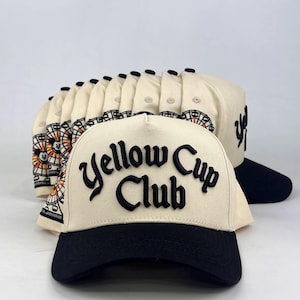 May include: A cream-colored trucker hat with a black brim and the text Yellow Cup Club embroidered in black on the front. The side of the hat features a graphic of a Ferris wheel with a small character face in the center, repeated across a stack.