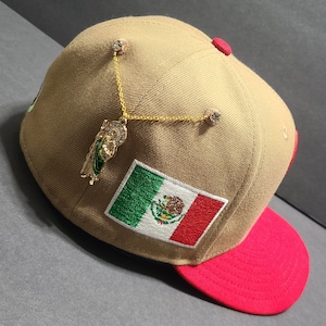May include: Tan baseball cap with a red brim and a Mexican flag patch. The cap is adorned with a gold chain and charm, and two rhinestone accents. The cap is angled, showing the details of the design.