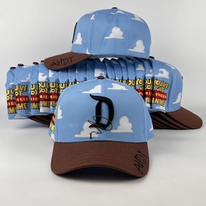 Könnte beinhalten: Eine hellblaue Baseballkappe mit weißem Wolkenmuster, einem braunen Schirm mit dem in Schwarz geschriebenen Namen Andy und einem großen schwarzen Buchstaben D auf der Vorderseite. An der Seite steht der Text You've Got A Friend In Me in gelben und roten Buchstaben.