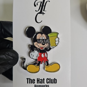May include: A Mickey Mouse enamel pin with sunglasses and a green drink. The pin is on a white card with "The Hat Club Accessories" printed at the bottom. The pin is black, red, yellow, and green.
