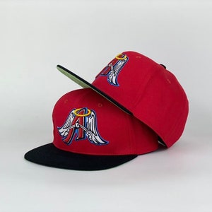May include: Two red baseball caps with a black brim and a green underside. Each cap features a stylized "A" with angel wings and a halo in blue, white, and gold embroidery. The caps are stacked on top of each other.