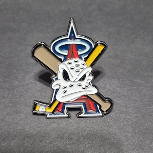 May include: A metal pin featuring the Anaheim Angels logo with a cartoon duck-like character. The pin has a silver border, with a white face, crossed baseball bats, and a hockey stick. The background is a dark gray.