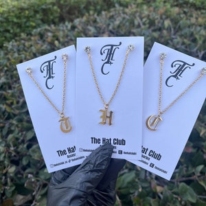 Könnte beinhalten: Goldfarbene Initialen-Halsketten und passende Ohrstecker werden auf weißen Karten präsentiert. Jede Halskette hat einen einzelnen Buchstabenanhänger, der an einer zarten Kette hängt. Die Karten tragen den Text "The Hat Club" und Social-Media-Handles.