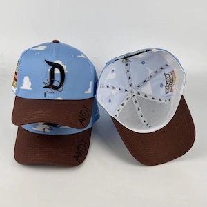 Könnte beinhalten: Eine hellblaue Baseballkappe mit weißem Wolkenmuster, braunem Schirm und einem schwarzen Buchstaben D auf der Vorderseite. Das Wort ANDY steht auf dem Schirm. Das Innere hat ein weißes Futter mit einem Logo-Muster und einer Toy Story-Grafik.
