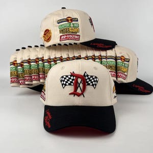May include: Cream-colored baseball caps with black brims. One cap features a red "D" with checkered flags, while others display racing-themed patches and text. The caps are stacked, showcasing their design.