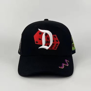 以下が含まれることがあります： 赤いサイコロのデザインと白い文字「D」が前面に刺繍された黒い野球帽。帽子には、紫と金のヘビのデザインやその他のカラフルなパッチも付いています。