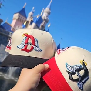 May include: Two baseball caps with cream-coloured crowns and black and red brims. One cap features a red "D" with angel wings, the other a black "P" with angel wings. Both have a gold halo above the design. The background shows a blue sky and a castle.