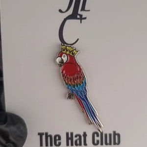 May include: A colorful parrot pin with a red body, blue and yellow tail feathers, and a gold crown. The pin is on a white card with the text "The Hat Club Accessories". The parrot pin is a unique accessory.