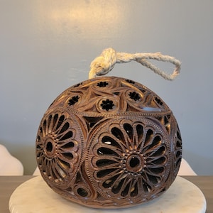 Op de afbeelding: Een bruine, ingewikkeld gesneden houten bol met bloemen- en geometrische patronen. Een natuurlijk touw is aan de bovenkant bevestigd. De bol wordt getoond op een wit marmerachtig oppervlak.