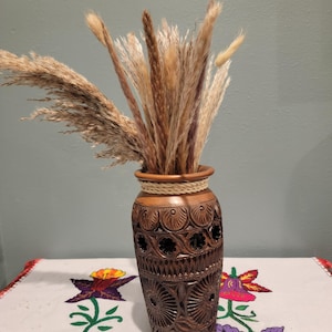 May include: A brown wooden vase with intricate carved designs, holding a bundle of dried, beige-colored ornamental grasses. The vase has a woven rope detail around the top. The vase sits on a white tablecloth with floral embroidery.