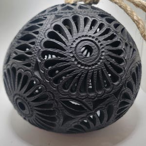 Op de afbeelding: Een zwart, bolvormig ornament met ingewikkelde bloemenpatronen. Een natuurlijke touwlus is aan de bovenkant bevestigd om op te hangen. Het oppervlak van het ornament heeft een textuur en het ontwerp laat licht door.