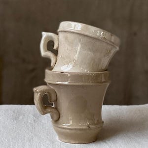 May include: Two stacked ceramic cups in a light beige, off-white tone. Each cup features a wide, flared rim and a distinctively shaped, angular handle. The surface of the cups has a smooth, glazed finish with subtle variations in color and texture.