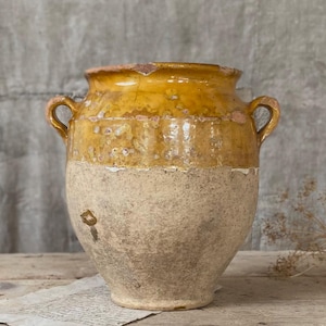 Può includere: Un vaso rustico in ceramica a due toni, con una parte superiore smaltata giallo-arancio e una metà inferiore beige strutturata. Il vaso ha due piccole maniglie e un bordo leggermente scheggiato. È appoggiato su una superficie di legno.