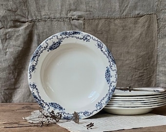 May include: A stack of white ceramic plates with a blue floral design around the rim. The plates are on a wooden surface with a neutral-colored fabric backdrop. A single plate is in the foreground, showcasing the intricate blue pattern.