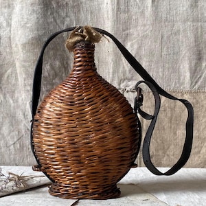 May include: A woven wicker bottle with a leather strap. The bottle is brown and has a natural finish. The strap is black and has a buckle.