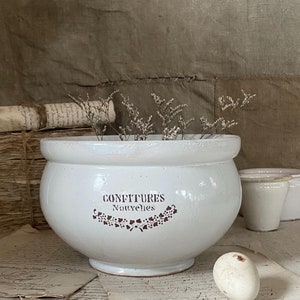 May include: White ceramic pot with a brown inscription that reads "CONFITURES Nouvelles". The pot is decorated with a brown floral design and is filled with white flowers.