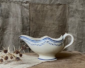 May include: A white ceramic gravy boat with a blue and white floral design. The boat has a handle and a spout. It is sitting on a wooden surface.