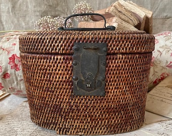 May include: Oval-shaped, woven wicker basket with a hinged lid and a metal clasp. The basket has a dark brown finish and a black metal handle. The clasp is rectangular and has a decorative design. The basket is likely used for storage.