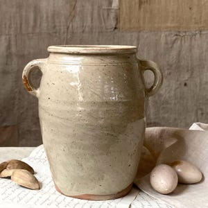 Puede incluir: Una vasija de cerámica beige con dos asas laterales y un acabado brillante. El recipiente presenta una boca ancha, un cuerpo ligeramente cónico y un moteado sutil en todo el esmalte. Dos objetos ovalados lisos y claros descansan cerca de la base.