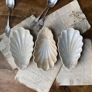 以下が含まれることがあります： 手書きのテキストが書かれたヴィンテージの本の上に、白いセラミック製の貝殻の形をした皿が3つ置かれています。皿はサイズと形がわずかに異なり、質感のある波打った表面をしています。