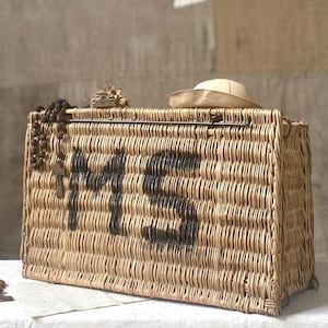 Puede incluir: Una cesta rectangular de mimbre trenzado con las letras MS pintadas en negro en la parte frontal. Un rosario de cuentas de color marrón oscuro con una cruz de metal cuelga del lado izquierdo y un sombrero de tela beige descansa sobre la tapa.