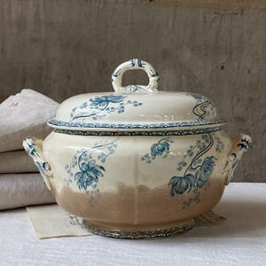 May include: A cream-coloured ceramic tureen with a matching lid, featuring blue floral patterns and a decorative border. The base of the tureen has a light brown gradient, and it includes two side handles and a central handle on the lid.