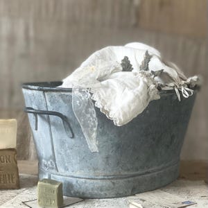 May include: A vintage galvanized metal wash tub filled with white linens. The tub has two handles and shows signs of wear. A bar of green soap and a stack of soap are in the foreground, with vintage ephemera.