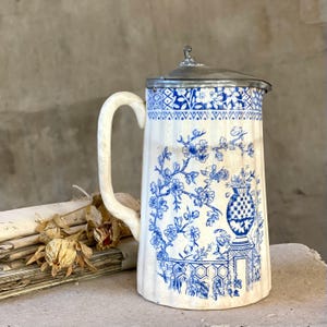 Puede incluir: Una jarra de cerámica blanca vintage con un diseño floral azul y una tapa de metal. La jarra tiene un asa curva y un patrón detallado de flores y un jarrón. El diseño recuerda a la porcelana clásica azul y blanca.