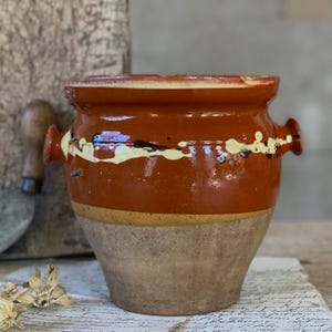 May include: A vintage ceramic pot with a reddish-brown glazed top and a lighter, unglazed base. The pot has two small handles and a decorative white and black painted band. This pot is likely for cooking or storage, and has a rustic, antique style.
