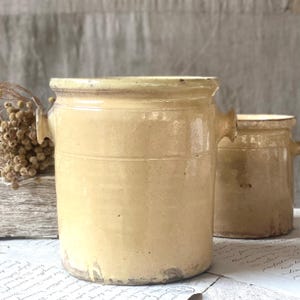 Può includere: Due antichi vasi in gres giallo chiaro con manici. Il vaso più grande ha un bordo leggermente svasato e due fasce orizzontali. Il vaso più piccolo è sullo sfondo. Entrambi hanno un aspetto rustico e invecchiato.