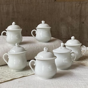 Pode incluir: Um conjunto de seis pequenas chávenas de porcelana branca com tampa, cada uma com um corpo arredondado, uma pega curva e uma tampa abobadada a condizer com um pequeno puxador. As chávenas têm um acabamento liso e brilhante.