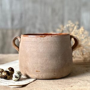 Puede incluir: Una rústica olla de cerámica beige con dos asas curvas. La olla tiene una superficie texturizada y un borde marrón rojizo. Varios huevos de codorniz moteados descansan sobre un trozo de papel envejecido.